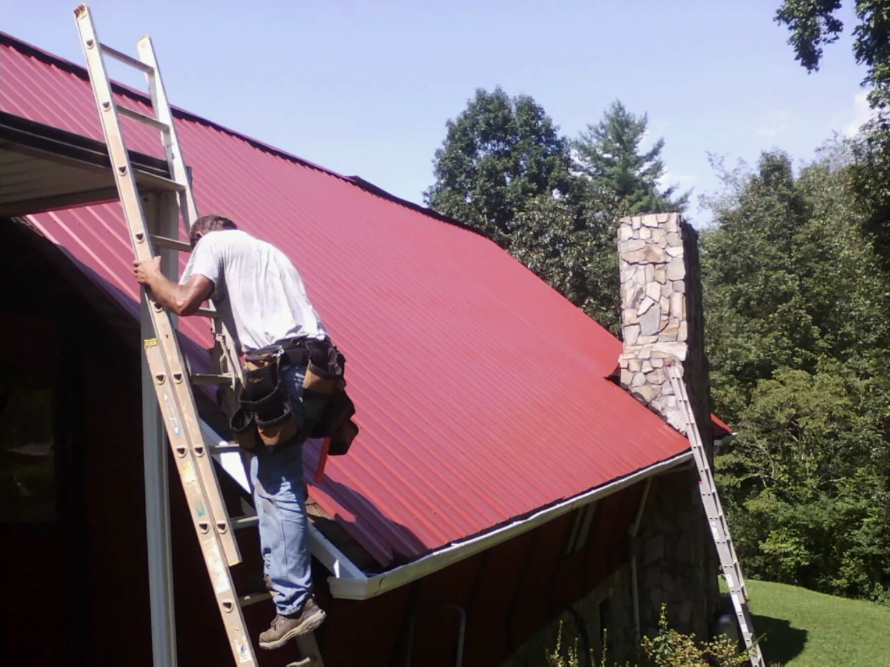 Roofer on a ladder beside a red metal roof and stone chimney — Soffit & Fascia Repair by Roofing Brothers in Harpers Ferry